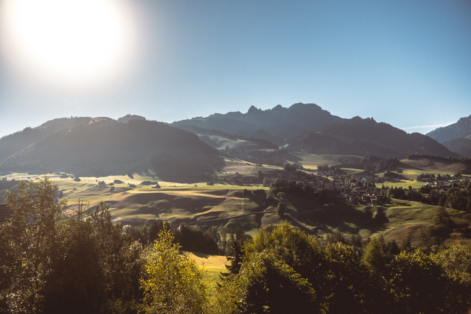 The Gruyere pre-Alps on the horizon in the haze of a summer morning, the sun flaring above the ridge, a village settled on the green slopes across the valley.