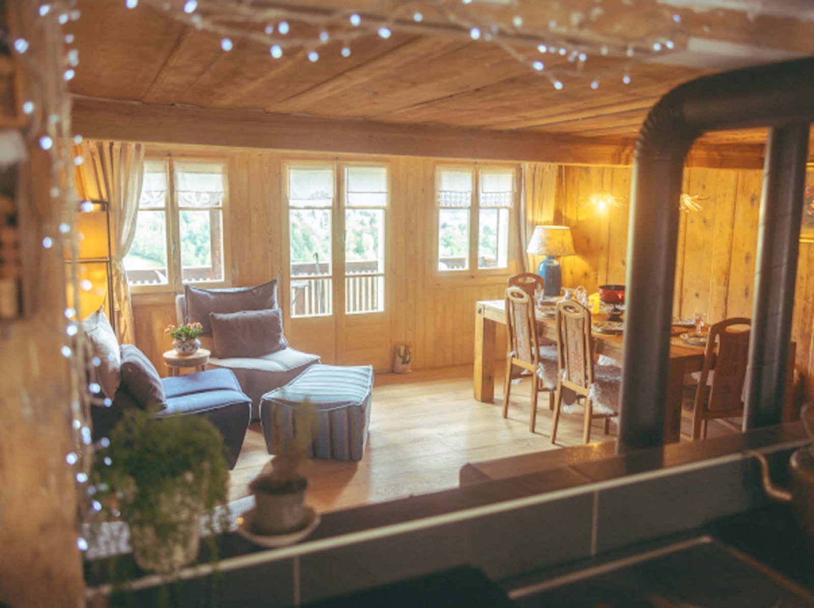 The open living and dining room under a ceiling of fairy lights, a soft blue sofa, the long table set for six, and the balcony doors framing the valley beyond.