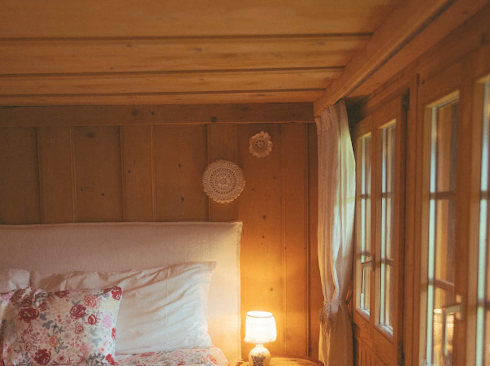 A bed dressed in white cotton and florals, a lamp glowing beside the balcony doors at dusk.